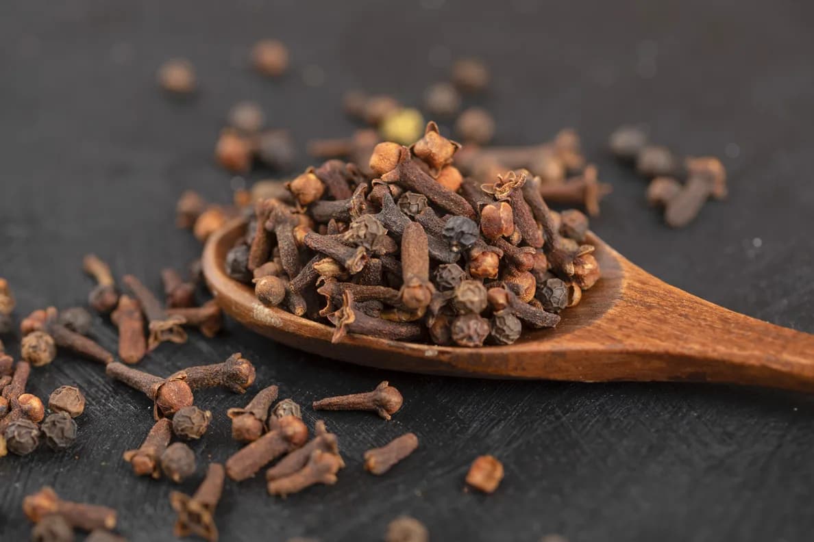 Ahşap bir kaşıkta kuru karanfil taneleri, koyu zeminde dağılmış karanfillerle birlikte